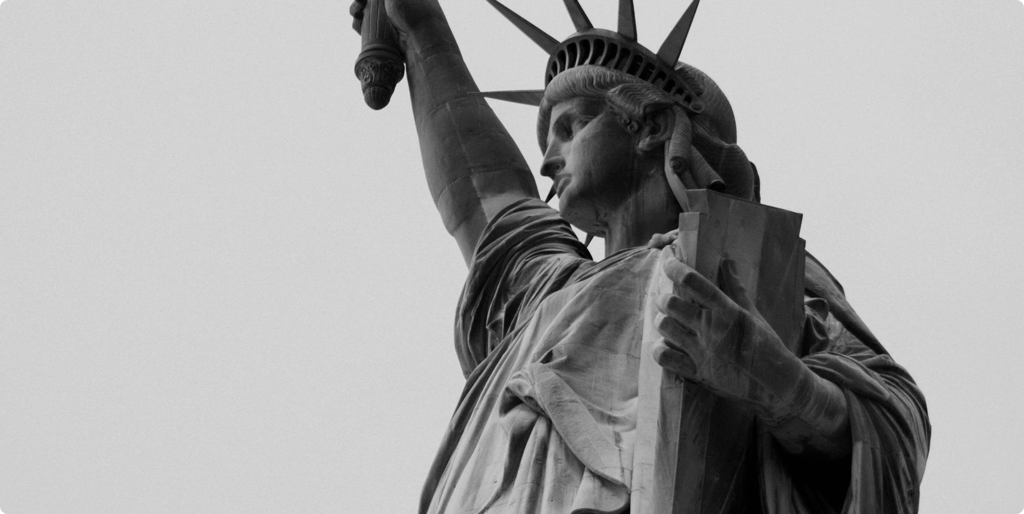 Close-up monochrome view of the Statue of Liberty’s upper torso and face, showing her raised torch arm, draped robes, and tablet held in her left hand against a plain sky.