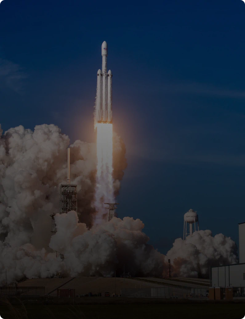 SpaceX Falcon Heavy rocket lifting off from its launch pad, bright plume of fire and smoke billowing against a deep blue sky.