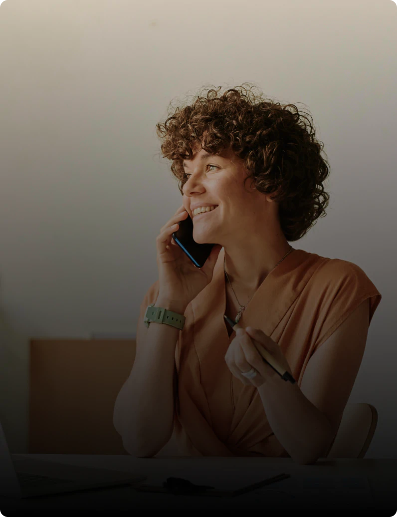 Smiling curly-haired woman holding a pen at her desk while talking on a smartphone, with an open laptop in front of her.
